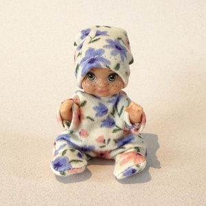 May include: A small doll wearing a matching hat and onesie. The doll's outfit is white with a pattern of blue and pink flowers. The doll has a smiling face with freckles and is sitting with its knees bent.
