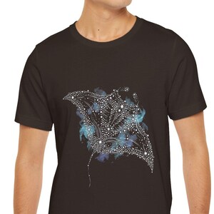 May include: A dark brown t-shirt featuring a detailed illustration of a manta ray. The design incorporates white dots and blue accents, creating a celestial or underwater theme. The t-shirt is made of a soft material.