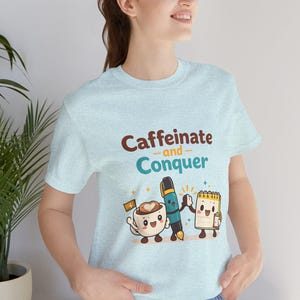 以下が含まれることがあります： 「Caffeinate and Conquer」の文字が茶色とティール色でプリントされた、水色のTシャツ。コーヒーカップ、ペン、メモ帳のキャラクターが描かれています。柔らかい素材のシャツです。