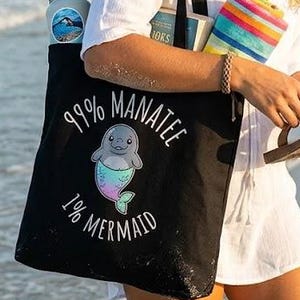 May include: A black tote bag with the text "99% Manatee 1% Mermaid" and a cartoon manatee with a mermaid tail. The bag is carried by a person on a beach, along with a water bottle, books, and a colourful striped towel.