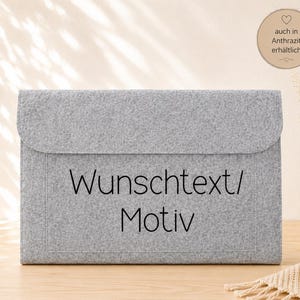 Könnte beinhalten: Eine hellgraue Dokumententasche aus Filz mit Klappverschluss. Auf der Vorderseite der Tasche befindet sich der schwarze Aufdruck "Wunschtext/ Motiv". Ein runder Aufkleber in der oberen rechten Ecke enthält den Text "auch in Anthrazit erhältlich".