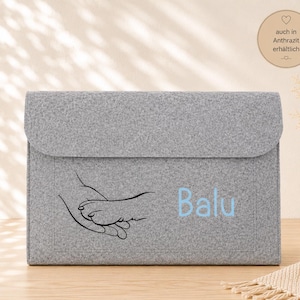 Könnte beinhalten: Eine hellgraue Dokumentenmappe aus Filz mit einer schwarzen Strichzeichnung einer menschlichen Hand, die eine Pfote hält. Der Name Balu ist in hellblauen Buchstaben auf der rechten Seite aufgedruckt. Die Mappe ist rechteckig und hat einen Klappverschluss.