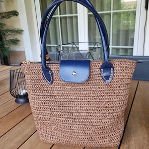 May include: A woven tote bag with a brown body and navy blue leather handles and accents. The bag features a navy blue flap closure and a gold zipper. The bag is sitting on a wooden surface.
