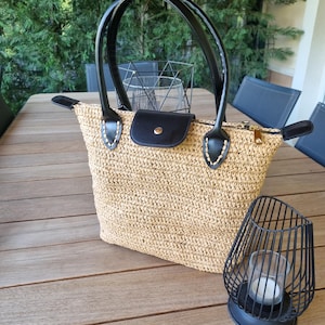 May include: A woven straw tote bag with black leather handles and accents. The bag features a black flap closure and a gold zipper. A black metal candle holder with a lit candle sits nearby. The bag is on a wooden table.
