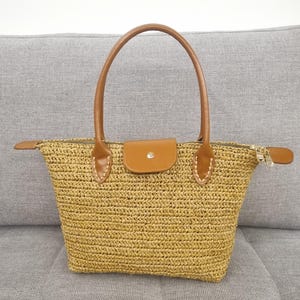 May include: A straw tote bag with brown leather handles and trim. The bag has a zipper closure and a small leather flap with a metal clasp.