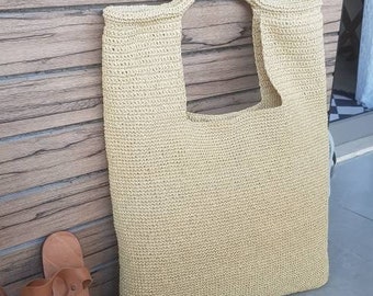 May include: A beige crocheted tote bag with a wide, open top and a single handle. The bag is sitting on a gray tiled floor next to a pair of brown sandals with colorful beaded embellishments.