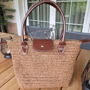 May include: A woven straw tote bag with brown leather handles and a matching flap closure. The bag features a zippered top and is designed for carrying items. The bag is sitting on a wooden surface.