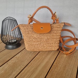 Op de afbeelding: Een klein, bruin strooien handtasje met een bruin leren klepje en een bruin leren bandje. De tas heeft een gouden rits en een gouden knoop.