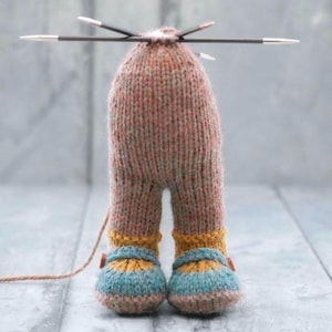 Op de afbeelding: Handgemaakte gebreide pop in bruin, geel en blauw. De pop heeft een afgeronde kop met breinaalden erin, benen en blauw-gele gestreepte schoenen. Een bruin garen is aan de pop bevestigd.