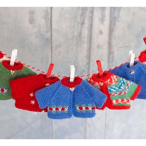 Puede incluir: Una línea de suéteres de punto en miniatura en varios colores y patrones, incluyendo rojo, verde, azul y blanco, colgados de una cuerda para tender la ropa con pinzas.