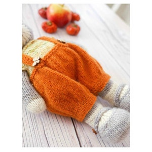 K&ouml;nnte beinhalten: Ein gestricktes Spielzeug mit orangefarbener Latzhose und grauen gestrickten Beinen und F&uuml;&szlig;en. Das Spielzeug liegt auf einer wei&szlig;en Holzoberfl&auml;che.