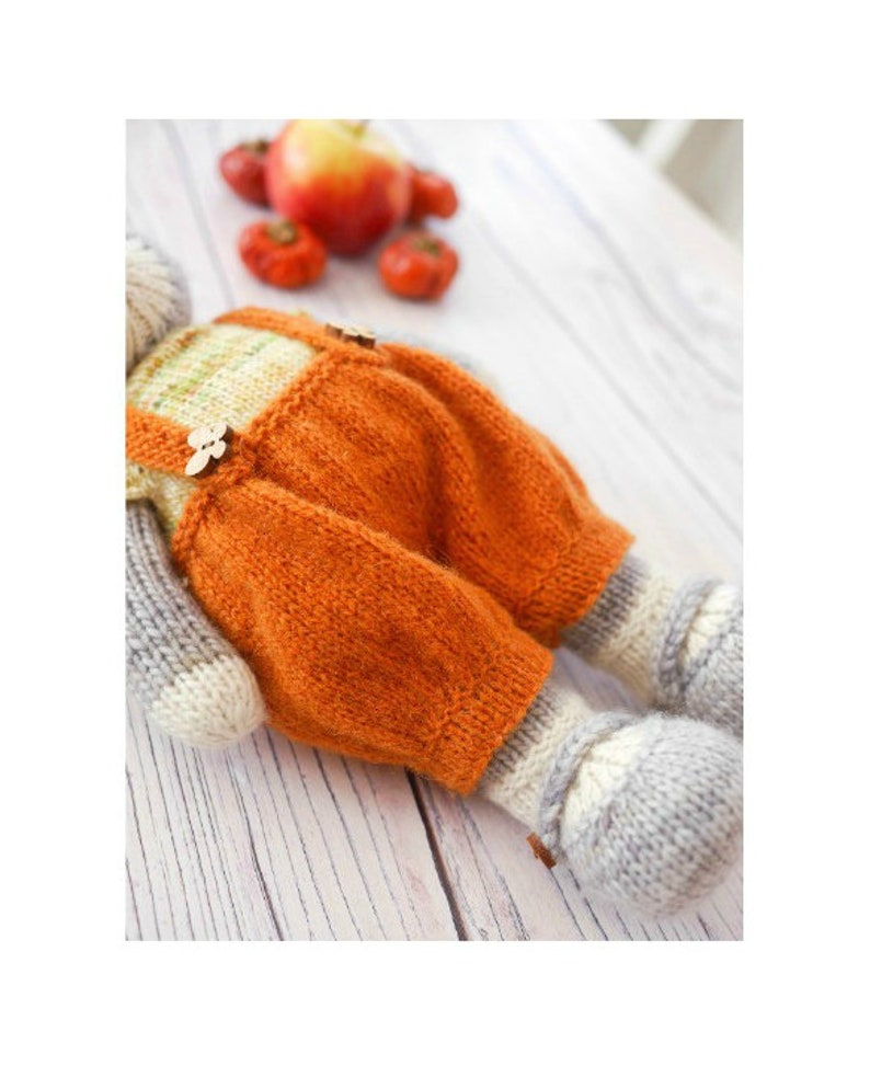 K&ouml;nnte beinhalten: Ein gestricktes Spielzeug mit orangefarbener Latzhose und grauen gestrickten Beinen und F&uuml;&szlig;en. Das Spielzeug liegt auf einer wei&szlig;en Holzoberfl&auml;che.