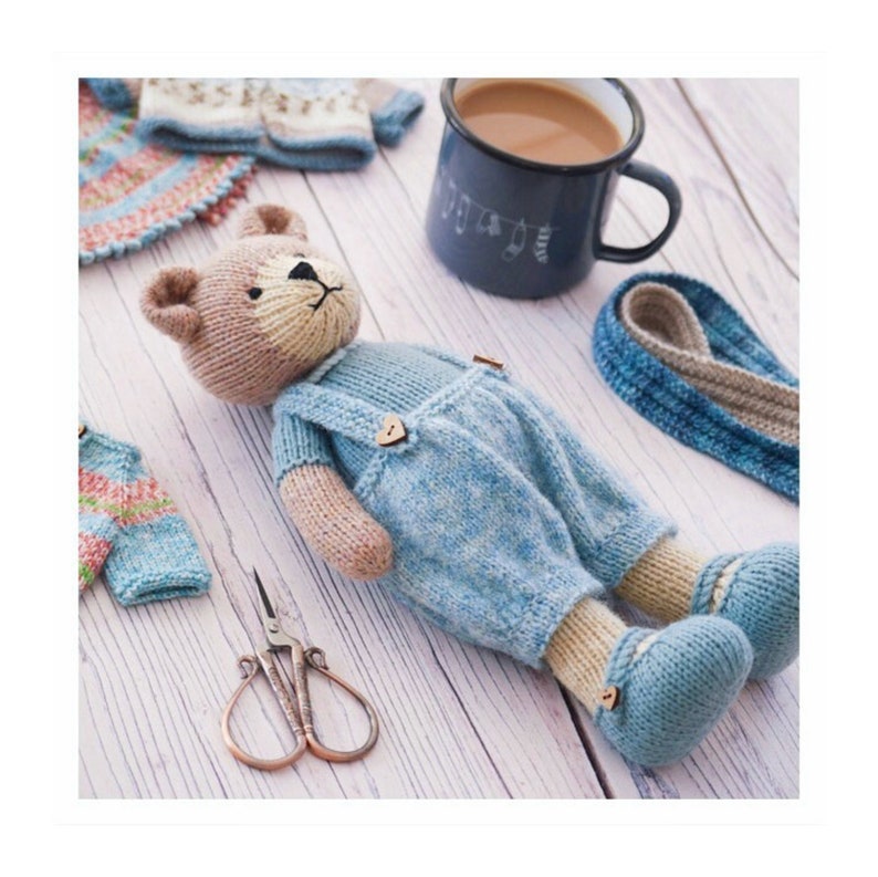 K&ouml;nnte beinhalten: Ein gestrickter Teddyb&auml;r, der eine blaue Latzhose mit Holzkn&ouml;pfen tr&auml;gt. Der B&auml;r liegt auf einer wei&szlig;en Holzoberfl&auml;che mit einer kleinen Schere und anderen Strickwaren.