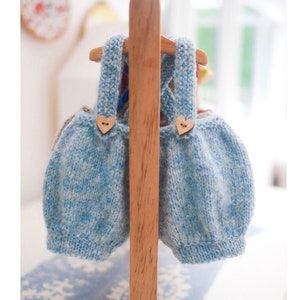 K&ouml;nnte beinhalten: Zwei blaue und wei&szlig;e gestrickte Baby-Latzhosen mit Holz-Herz-Kn&ouml;pfen, die an einem Holzgestell h&auml;ngen.