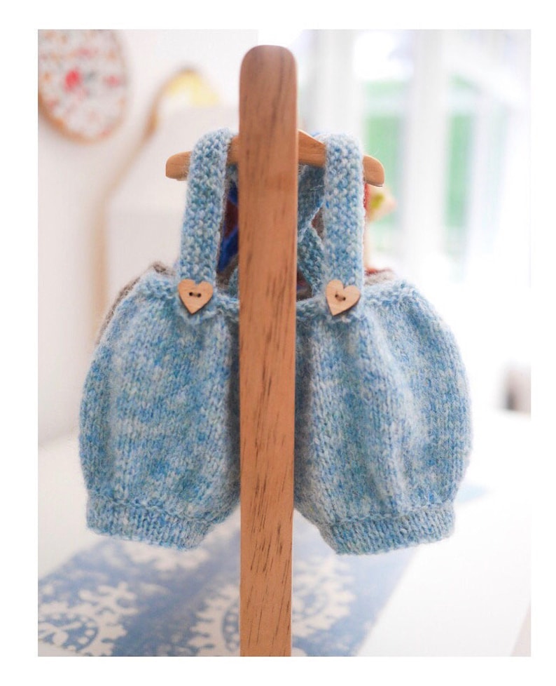 K&ouml;nnte beinhalten: Zwei blaue und wei&szlig;e gestrickte Baby-Latzhosen mit Holz-Herz-Kn&ouml;pfen, die an einem Holzgestell h&auml;ngen.