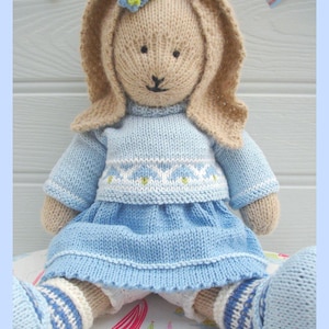 Peut inclure: Une poupée lapin tricotée à la main de couleur beige avec de longues oreilles, portant un pull bleu clair avec une bande à motifs blancs et une jupe bleue assortie. Le lapin a deux petites fleurs bleues sur la tête et des chaussettes rayées bleu et blanc. Texte : COPYRIGHT SUSAN HICKSON 2009.
