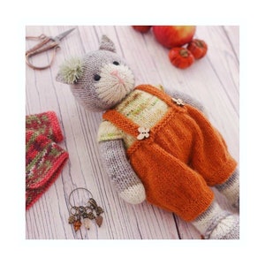 K&ouml;nnte beinhalten: Ein graues, gestricktes Katzenspielzeug, das eine orange Latzhose mit Holz-Kn&ouml;pfen tr&auml;gt. Das Spielzeug sitzt auf einer wei&szlig;en Holzoberfl&auml;che.