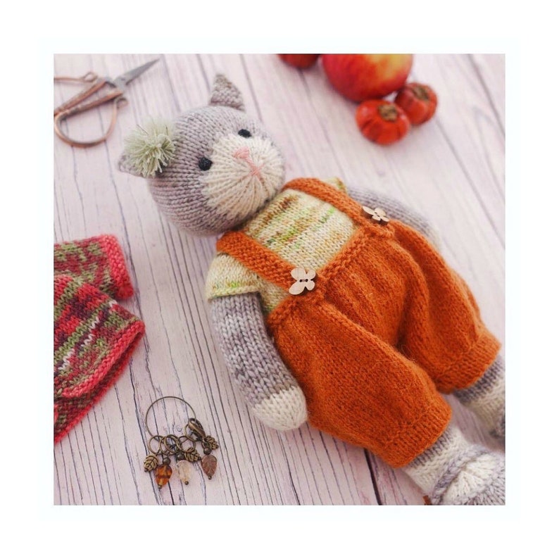 K&ouml;nnte beinhalten: Ein graues, gestricktes Katzenspielzeug, das eine orange Latzhose mit Holz-Kn&ouml;pfen tr&auml;gt. Das Spielzeug sitzt auf einer wei&szlig;en Holzoberfl&auml;che.