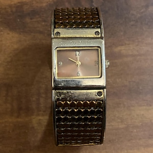 May include: A rectangular wristwatch with a gold-tone case and a brown, textured band. The watch face is a light pink color with gold-tone hands and hour markers. The band appears to be made of small, connected metal pieces.