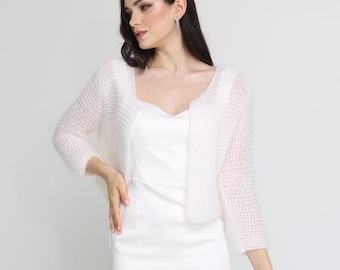 May include: A white, floor-length gown with a matching, open-front, short-sleeved bolero. The bolero is a delicate, open-weave knit. The model is wearing gold-coloured sandals. The background is a solid white.