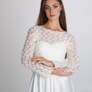 May include: A white lace long-sleeved top with a boat neckline. The top is paired with a white satin skirt. The intricate lace detailing adds a touch of elegance to the bridal or formal wear.