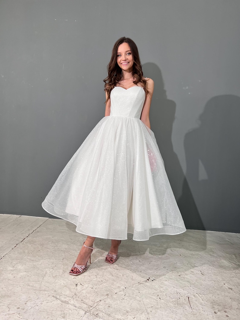 May include: A white tulle midi dress with sweetheart neckline and subtle glitter details. The A-line skirt adds volume and creates a flattering silhouette. The dress is shown on a model against a gray background.