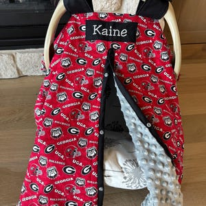May include: A red and white baby car seat cover featuring the University of Georgia Bulldogs logo and the name "Kaine". The cover has a black trim and a soft, gray interior. The car seat is made by Graco.