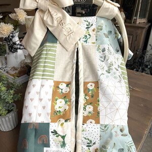 May include: A patchwork baby car seat cover with a large cream-colored bow and the name "Paisley" embroidered on a cream-colored tag. The cover features various fabric patterns, including floral, geometric, and rainbow designs, in shades of green, brown, and cream.