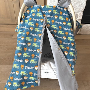 May include: A teal baby car seat cover featuring a repeating pattern of Scooby-Doo characters and vans. The cover has a gray trim and a button closure. The car seat handle is gray with the brand name "GRACO" printed on it.