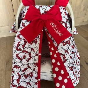 May include: A baby car seat cover featuring a white background with a repeating pattern of Hello Kitty faces with red bows. The cover has a large red bow and a red polka dot flap. The name "Basani" is embroidered on the bow.