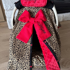 May include: A baby car seat cover with a leopard print design. The cover features a large red bow and a red headrest with the name "Oaklynn" embroidered in black. The edges are trimmed in black and red, with red buttons.