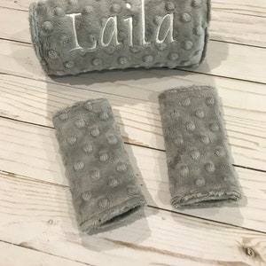 May include: Gray, plush baby car seat accessories. The set includes a headrest with the name "Laila" embroidered in white, and two strap covers. The fabric has a textured, dotted pattern.