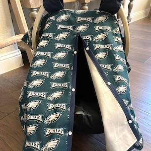 May include: A dark teal baby car seat cover featuring the Philadelphia Eagles logo in white. The cover has a navy blue trim and white snap closures. The car seat handle is visible at the top, with the brand name "GRACO" printed on it.