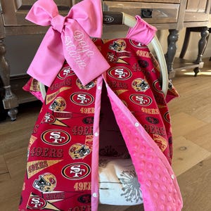 May include: A red and gold baby car seat cover featuring the San Francisco 49ers logo and helmet design. It has a pink bow with "Deya Marie" embroidered on it. The cover is lined with pink fabric and has white buttons.