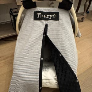May include: A baby car seat cover with a black and white striped pattern. The cover has a black trim and a black panel with a textured pattern. The name "Tharpe" is embroidered on a black patch. The car seat handle has the brand name "Graco".