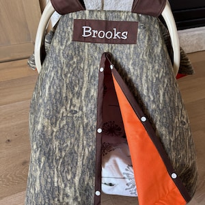 May include: A camouflage-patterned baby car seat cover with the name "Brooks" embroidered on a brown patch. The cover has an orange interior and is trimmed with brown fabric. The Graco logo is visible on the handle.