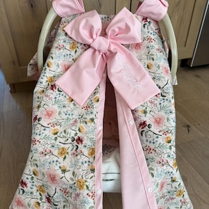 May include: A floral patterned baby car seat cover with a large pink bow and the name "Annie" embroidered on it. The cover has a light pink trim and is secured with buttons. The car seat frame is visible at the top.