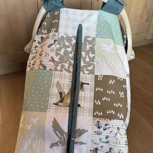 May include: A patchwork baby car seat cover featuring a duck theme. The cover is made of various fabric squares in shades of green, brown, and white, with images of ducks, geese, and trees. A teal fabric strip with snap closures runs down the center.