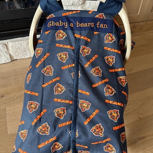 May include: Navy blue baby car seat cover featuring a repeating pattern of orange Chicago Bears logos and text. The cover has a blue trim and a banner that reads "Gbaby a bears fan" in orange thread. The cover is designed to fit a Graco car seat.