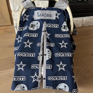 May include: A navy blue baby car seat cover featuring a repeating pattern of white stars, football helmets, and the word "Cowboys." The cover has a gray trim and the name "Lucas" embroidered on it. The car seat is made by Graco.