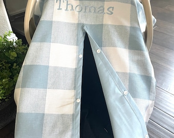 Peut inclure: Une housse de siège auto pour bébé à carreaux bleus et blancs. Le nom "Thomas" est brodé en bleu clair. La housse se ferme avec des boutons et possède une bordure bleu clair. La marque "LilCarsonBlue" est visible.