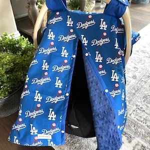 May include: Blue and white baby car seat cover featuring the Los Angeles Dodgers logo and baseballs. The cover has a soft, textured interior and attaches to the car seat with snaps. The cover is designed to fit a Graco car seat.