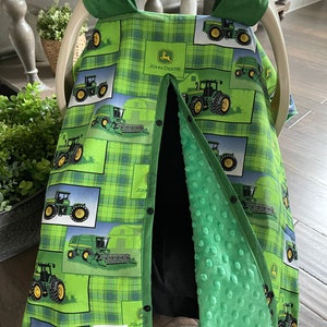 May include: Green and blue plaid baby car seat cover with tractor graphics and the John Deere logo. The cover has a black interior and a green, textured lining. The cover is secured with black buttons.