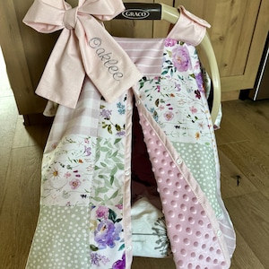 May include: A baby car seat cover with a large pink bow and the name "Oaklee" embroidered on a strap. The cover features a patchwork design with floral and polka dot patterns in shades of pink, green, and white.