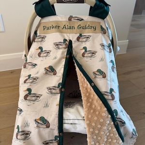 May include: A baby car seat cover with a duck pattern. The cover is cream-colored with illustrations of ducks swimming in water. The trim is dark green, and the interior is a soft, tan fabric. The name "Parker Alan Guidry" is printed on the top.