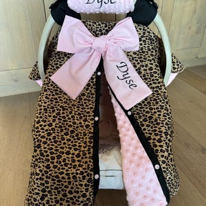 May include: A baby car seat cover with a leopard print design. The cover features a large pink bow, a pink headrest with the name "Dyse" embroidered in black, and a pink, dimpled interior lining. Black trim and white buttons complete the design.