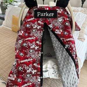 May include: A red and white patterned baby car seat cover with the word "Parker" embroidered on a black patch. The cover features the word "GEORGIA" and the Georgia Bulldogs logo. The interior is gray and textured.