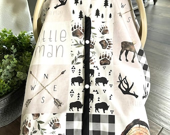 Peut inclure: Une housse de siège auto pour bébé avec un motif patchwork comprenant des empreintes de pattes d'ours, des silhouettes de bisons, des arbres et des flèches de boussole. La housse a un motif à carreaux noir et blanc et le texte "Little Man".