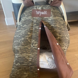 Puede incluir: Una funda para asiento de coche de beb&eacute; con un estampado de camuflaje en tonos marrones y grises. La funda tiene un ribete marr&oacute;n y el nombre "Wyatt" bordado en un parche marr&oacute;n. La funda est&aacute; parcialmente abierta, revelando un interior con estampado floral.