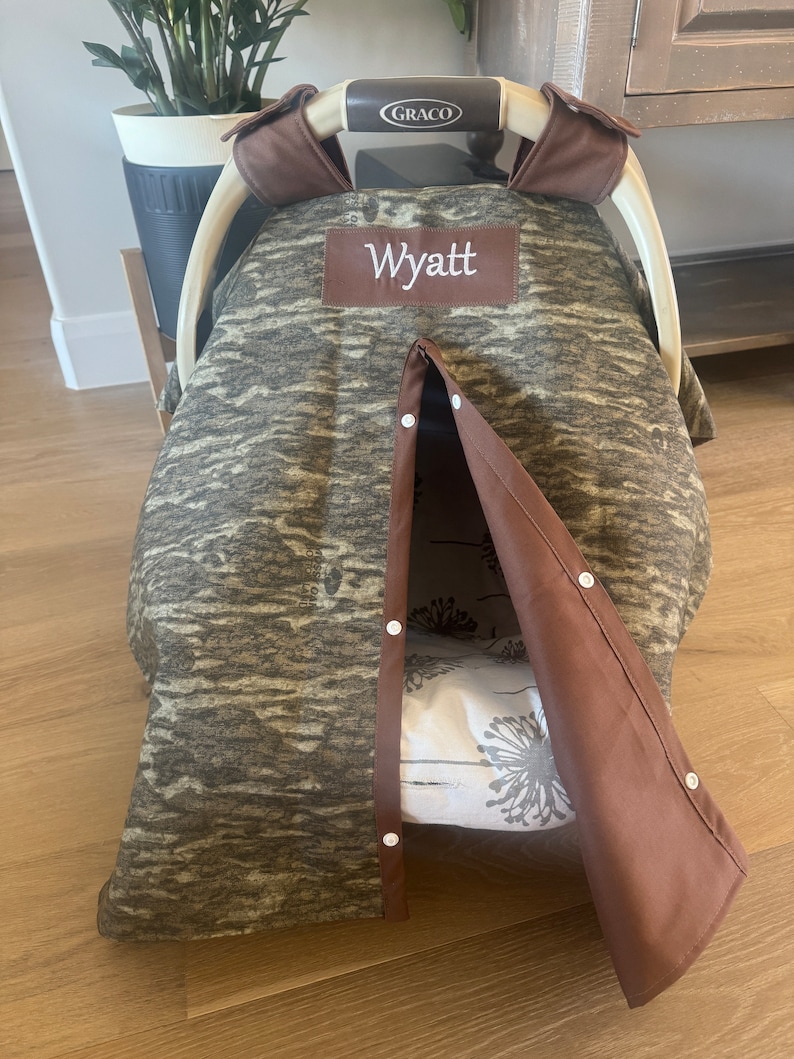 Puede incluir: Una funda para asiento de coche de beb&eacute; con un estampado de camuflaje en tonos marrones y grises. La funda tiene un ribete marr&oacute;n y el nombre "Wyatt" bordado en un parche marr&oacute;n. La funda est&aacute; parcialmente abierta, revelando un interior con estampado floral.